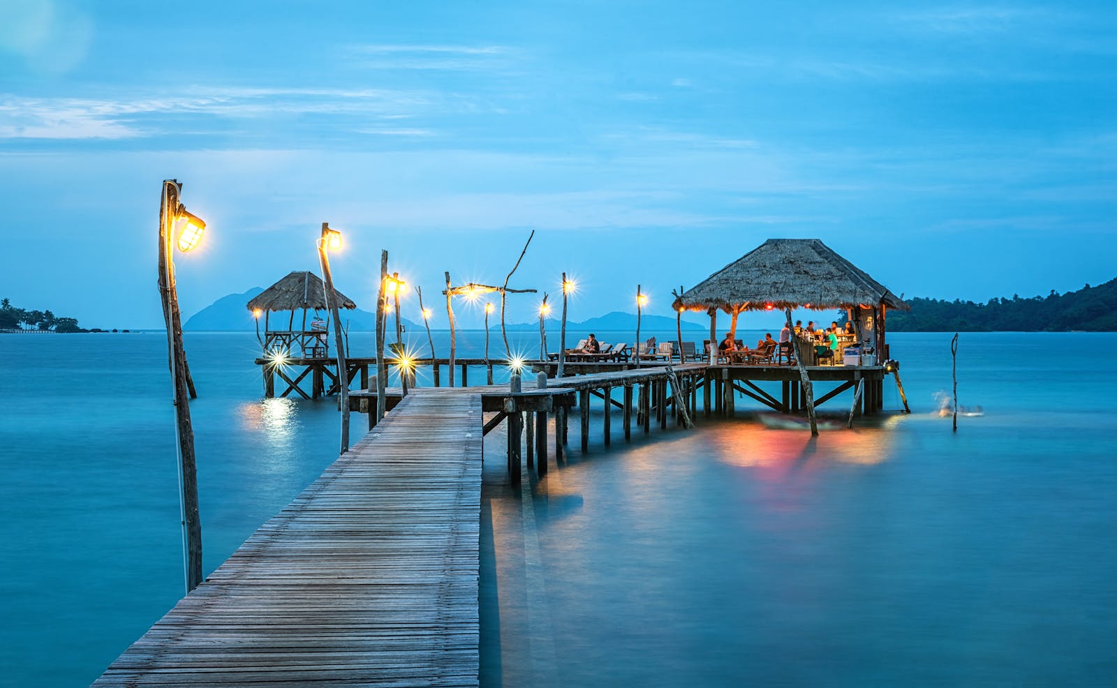 Aerial view of a Caribbean beach resort featuring a turquoise swimming pool, white sand beach, tall palm trees, thatched-roof buildings, a wooden pier, and neatly arranged rows of white lounge chairs under warm tropical sunlight.